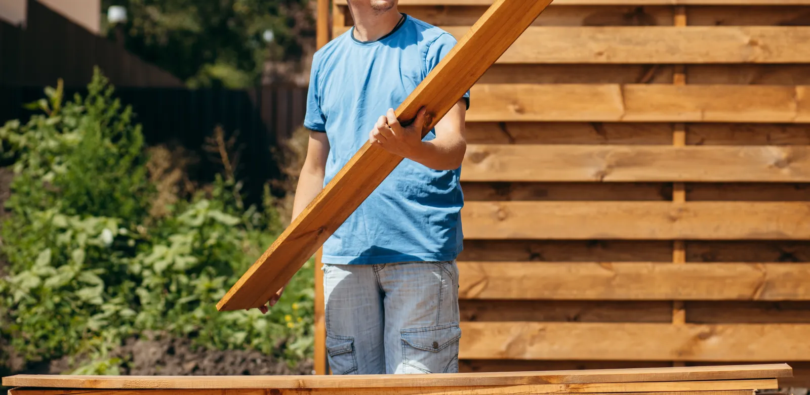 A fence installer carrying a cedar board during a horizontal wood fence installation in a residential backyard.